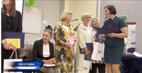 Kilka kobiet stoi na środku sali i jedna siedzi na wózku inwalidzkim - w rękach trzymają upominki i gratulacje, które jedna z widocznych kobiet im rozdaje