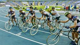 Pokaż zdjęcie: Sześciu parasportowców stoi na bieżni. W rękach trzymają rowery. Wyścig się skończył. Jedna z osób trzyma w rękach kwiaty. W tle trybuny z kibicami.