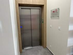 Pokaż zdjęcie: Winda z metalowymi rozsuwanymi drzwiami. Do dokoła obrobiona ramą z drewna. Po prawej stronie tabliczka  z informacją o dofinansowaniu ze środków PFRON.