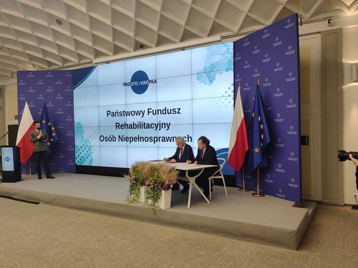 Pokaż zdjęcie: Sala konferencyjna z telebimem. U góry wyświetlone kolorowe logo Prosto i kropka, a poniżej tekst Państwowy Fundusz Rehabilitacji Osób Niepełnosprawnych. Po obu stronach telebimu flagi Rzeczpospolitej Polskiej i Unii Europejskiej. Z prawej strony stolik przy którym siedzą dwaj mężczyźni podpisujący dokumenty. Z lewej strony konferansjer z mikrofonem. Scena z ustawionymi w wazonach kwiatami.