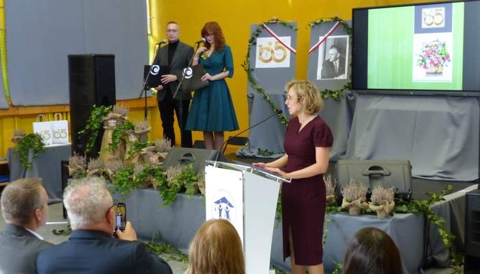 Pokaż zdjęcie: Kobieta w bordowej sukience stoi przy mównicy i odczytuje list gratulacyjny. Na scenie, przy mikrofonach ustawionych na statywach stoją kobieta wraz z mężczyzną. Scena udekorowana jest różnorodną roślinnością, w tym między innymi wrzosami.