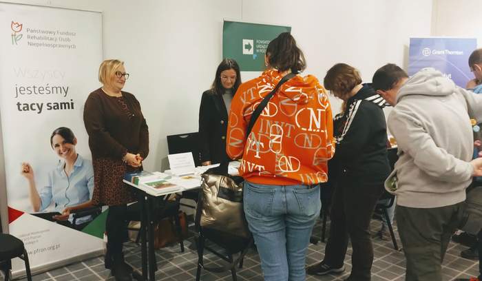 Pokaż zdjęcie: Dwie kobiety udzielają informacji osobom, które znajdują się przy stoisku Oddziału Wielkopolskiego PFRON. Na stoliku ułożone są materiały informacyjne. W tle ustawiony jest baner PFRON oraz banery wystawców sąsiadujących ze stoiskiem Oddziału.