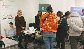 Pokaż zdjęcie: Dwie kobiety udzielają informacji osobom, które znajdują się przy stoisku Oddziału Wielkopolskiego PFRON. Na stoliku ułożone są materiały informacyjne. W tle ustawiony jest baner PFRON oraz banery wystawców sąsiadujących ze stoiskiem Oddziału.