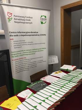 Pokaż zdjęcie: Na stołach przykrytych czerwonym płótnem znajdują się teczki z materiałami konferencyjnymi oraz różne ulotki. Za stolikami ustawiony jest baner CIDON.