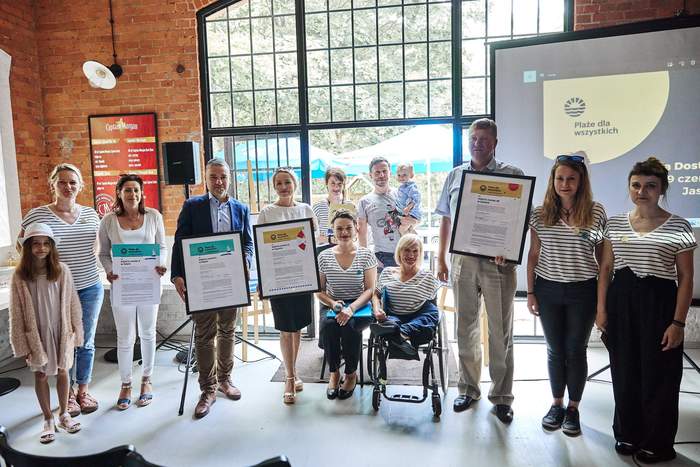 Pokaż zdjęcie: W rządzie stoi kilka osób niektóre z nich trzymają duże certyfikaty oprawione w ramkę. Po środku siedzi kobieta na wózku inwalidzkim. Wszyscy uśmiechają się do zdjęcia. W tle wielkie szklane loftowe drzwi i ściany z cegieł.