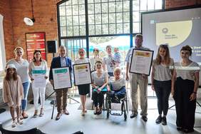 Pokaż zdjęcie: W rządzie stoi kilka osób niektóre z nich trzymają duże certyfikaty oprawione w ramkę. Po środku siedzi kobieta na wózku inwalidzkim. Wszyscy uśmiechają się do zdjęcia. W tle wielkie szklane loftowe drzwi i ściany z cegieł.
