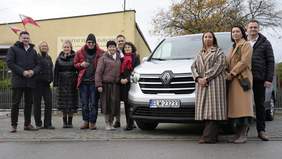 Pokaż zdjęcie: Przed budynkiem Warsztatu Terapii Zajęciowej znajduje się nowy samochód. Po jego prawej i lewej stronie stoją ludzie. Jest ich dziesięcioro, są to kobiety i mężczyźni. 