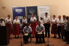 Pokaż zdjęcie: Na scenie siedzą dwaj mężczyźni, którzy grają na bębnach. Za nimi stoją kobiety ubrane w białe bluzki i czerwone spódnice oraz dwaj mężczyźni ubrani w białe koszule i czarne spodnie. W ręku maja małe instrumenty.