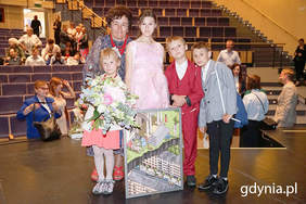 Pokaż zdjęcie: Kobieta w krótkich włosach, ubrana w kwiecistą sukienkę stoi z dwiema dziewczynkami i dwoma chłopcami. Wszyscy są galowo ubrani. Wyższa dziewczynka trzyma oparty na podłodze obraz, niższa trzyma kwiaty.