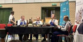 Pokaż zdjęcie: Grupa muzyczna Remont Pomp w niebieskich ogrodniczkach siedzi na scenie przy stole z instrumentami perkusyjnymi i przedmiotami codziennego użytku, tworzącymi zestaw do grania. W tle baner Radia Gdańsk oraz plakat plebiscytu „Lodołamacze”.