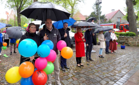 Grupa osób stojących na brukowanym placu podczas deszczu, chroniących się pod czarnymi i niebieskimi parasolami. Na pierwszym planie dwoje ludzi trzyma duży pęk kolorowych balonów (niebieskie, różowe, pomarańczowe, zielone). W tle widać zaparkowaną furgonetkę, wóz strażacki oraz osobę przebraną za klauna. Wszyscy są ubrani w odzież wierzchnią dostosowaną do pogody.