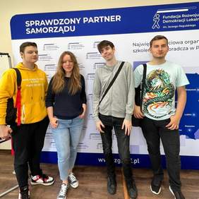 Pokaż zdjęcie: 3 uczniów oraz nauczycielka stoją obok siebie na tle banerów Fundacji Rozwoju Demokracji Lokalnej