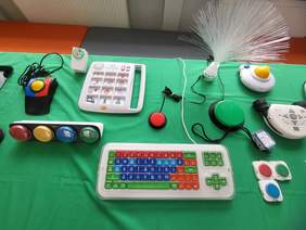 Pokaż zdjęcie: Na stole leżą różne sprzęty rehabilitacyjne i specjalistyczne narzędzia edukacyjne. Stół jest przykryty zielonym obrusem. 