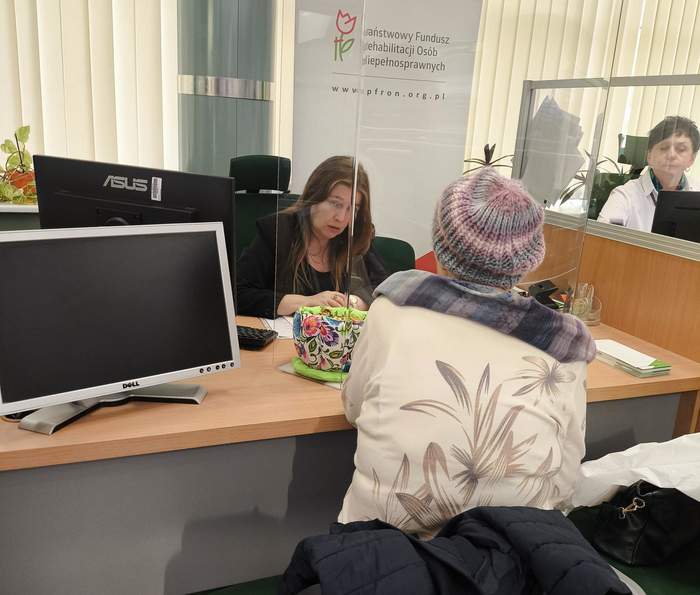 Pokaż zdjęcie: Przy biurku siedzą dwie kobiety naprzeciwko siebie, w tle stoi rollup z logo PFRON.