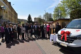 Pokaż zdjęcie: Grupa osób stoi na placu podczas uroczystości przekazania samochodu dostawczego Caritas Diecezji Drohiczyńskiej. W centrum duchowny odczytuje dokument, obok niego stoi duchowny oraz kobieta w różowej marynarce. Po prawej stronie widoczny biały samochód oznaczony logotypem Caritas i symbolem dostępności dla osób niepełnosprawnych, ozdobiony dużą czerwoną kokardą. W tle budynki mieszkalne i drzewa.