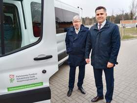 Obok samochodu stoi dwóch mężczyzn, którzy są bardzo elegancko ubrani. Na pojeździe naklejona jest naklejka Państwowego Funduszu Rehabilitacji Osób Niepełnosprawnych. 