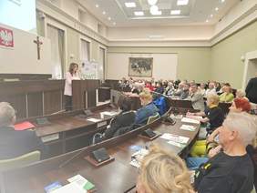 Pokaż zdjęcie: kobieta stoi na mównicy, przed nią słuchacze zasiadający na sali obrad w Urzędzie Miasta w Kielcach
