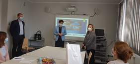 Pokaż zdjęcie: Anna Skupień, dyrektor Oddziału Wielkopolskiego PFRON ubrana w błękitną marynarkę przedstawia uczestnikom szkolenia jego prowadzących. Wszyscy mają założone na twarzy maseczki chroniące przed zakażeniem koronawirusem. W tle widoczny jest ekran, na którym wyświetlona jest prezentacja przedstawiająca funkcjonujące w Polsce Ośrodki Rehabilitacji Kompleksowej.