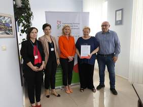 Pokaż zdjęcie: Na tle rollapu z logo PFRON pozuje pięć osób, jedna z kobiet trzyma w ręku podpisaną umowę.
