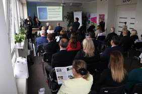 Pokaż zdjęcie: Pełna sala gości, wszyscy siedzą i słuchają kobiety, która omawia prezentację. Wszyscy są w maseczkach. 