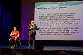 Pokaż zdjęcie: Na scenie jest dwóch mężczyzn w okularach jeden siedzi drugi trzyma w ręku mikrofon, w tle jest duży ekran z wyświetloną prezentacją