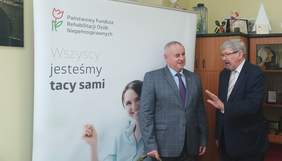 Pokaż zdjęcie: pomieszczenie w Oddziale Łódzkim PFRON, z tyłu widzimy baner z logo PFRON przy którym stoją uczestnicy spotkania pełnomocnik ds osób niepełnosprawnych Paweł Wdówik i Dyrektor Oddziału Łódzkiego Władysław Skwarka