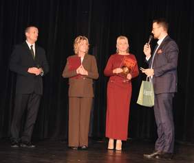 Czworo dorosłych osób stoi na scenie. Trzy z nich są ubrane formalnie (dwa garnitury, jeden damski komplet), a kobieta w czerwonej sukni trzyma w ręku ramkę lub dyplom. Mężczyzna po prawej, trzymający mikrofon, przemawia do zgromadzonych. Tło stanowi czarna, gładka kurtyna.