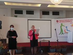 dwie panie stoją na jasnym tle, ta z lewej ubrana na czarno, druga w czarnej sukience i różowej marynarce mówi do mikrofonu