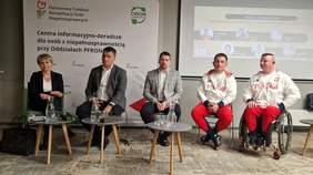 Pokaż zdjęcie: Paneliści kobieta i mężczyźni siedzą na środku sali na krzesłach. Dyskusję moderuje niewidomy mężczyzna - Marcin Ryszka. Wśród panleistów są osoby ubrane w biało-czerwone stroje sportowe.