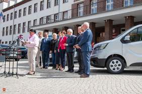 Pokaż zdjęcie: Uroczystość przed budynkiem placówki. Na pierwszym planie kobieta przemawia do mikrofonów ustawionych na statywach. Za nią stoi grupa ośmiu osób, a po prawej stronie widać nowy, srebrny bus, który jest przekazywany jednostce.