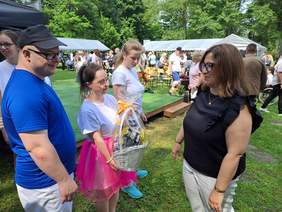 Pokaż zdjęcie: Podczas pikniku, kobieta przekazuje dzieciom kosz ze słodyczami. W tle widać namioty i uczestników pikniku.