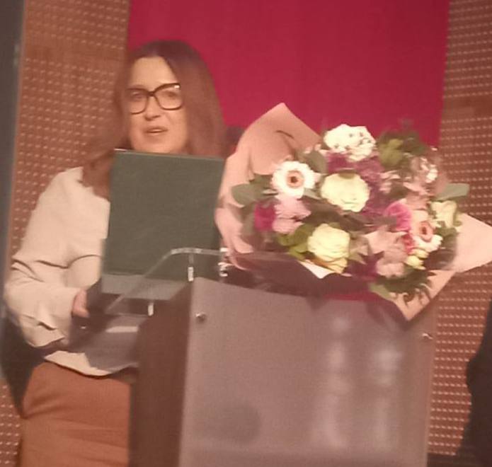 Pokaż zdjęcie: Przy mównicy na której leżą kwiaty, stoi kobieta. W ręku trzyma grawerton.