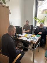 Przy biurku siedzi mężczyzna rozmawiający z dwoma pracownikami podczas dyżuru eksperckiego PFRON. Na stole znajdują się materiały informacyjne i komputer. Spotkanie odbywa się w jasnym pomieszczeniu biurowym z dużym oknem i rośliną w tle.