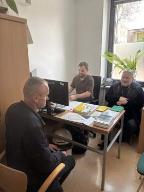 Przy biurku siedzi mężczyzna rozmawiający z dwoma pracownikami podczas dyżuru eksperckiego PFRON. Na stole znajdują się materiały informacyjne i komputer. Spotkanie odbywa się w jasnym pomieszczeniu biurowym z dużym oknem i rośliną w tle.