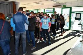 Pokaż zdjęcie: Fotografia przedstawia kolejkę osób do recepcji, gdzie odbywa się rejestracja uczestników koncertu. Osoby w kolejce (około 20 osób w pomieszczeniu, reszta osób czeka przed wejściem) mają na twarzach założone maseczki zgodnie z zaleceniami przeciwdziałającymi rozprzestrzenianiu się COVID-19 