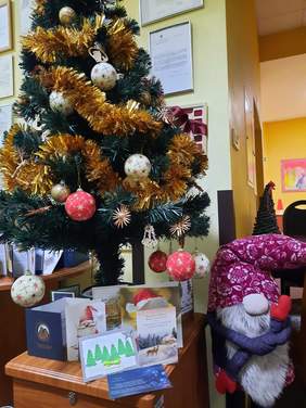 Pokaż zdjęcie: Po lewej stronie stoi choinka. Jest przystrojona bombkami, łańcuchami i ozdobami świątecznymi. Pod choinką leżą nadesłane kartki z życzeniami świątecznymi. Po prawej stronie stoi świąteczny skrzat. 