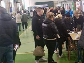 Pokaż zdjęcie: Tłum osób wewnątrz hali sportowej, ludzie w kurtkach stoją przy stolikach z ulotkami, jedna kobieta trzyma dziecko na ręku, w tle widoczne banery informacyjne i składane krzesła przy stanowiskach.