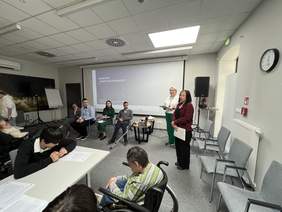 Pokaż zdjęcie: Prelegenci prowadzą warsztaty "Prawo do Dostępności" dla publiczności siedzącej przy stołach i na wózkach inwalidzkich w sali konferencyjnej.