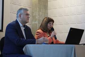 Pokaż zdjęcie: Przy stole konferencyjnym na którym stoi laptop siedzą dwie osoby. Od lewej mężczyzna w garniturze, po prawj kobieta szatynka w pomarańczowej marynarce przemawia doi uczestników konferencji 