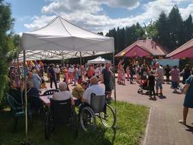 Pokaż zdjęcie: Pod rozstawionym namiotem przy stolikach siedzą seniorzy i osoby na wózkach. Przy scenie, na której podskakuje animatorka, gromadzi się tłum dorosłych z dziećmi. W dali widać dmuchaną zjeżdżalnię. Scenę przyozdabiają balony i ogromny obraz z