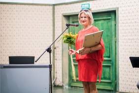 Pokaż zdjęcie: Zdjęcie z gali. Przy mikrofonie pani w pomarańczowej sukience, trzymająca kwiat słonecznika i oprawiony w ramę dyplom