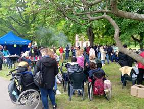 Pokaż zdjęcie: Na świeżym powietrzu piknikuje duża grupa osób, w tle namioty, na pierwszym planie osoby na wózkach inwalidzkich.