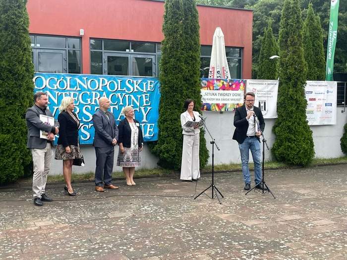Pokaż zdjęcie: Grupa osób stoi na zewnątrz podczas oficjalnego wydarzenia. Za nimi widoczny jest niebieski baner z napisem „Ogólnopolskie Integracyjne Warsztaty Artystyczne”. Na scenie, przed mikrofonem, przemawia mężczyzna w okularach i czarnej marynarce.