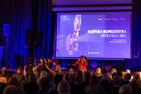 Pokaż zdjęcie: Zdjęcie zrobione podczas wydarzenia, na którym na scenie widać grupkę osób przygotowujących się lub występujących. Tło stanowi duży ekran z projekcją graficzną, na której widnieje napis: "RAZEM DLA BEZPIECZEŃSTWA - PREWENCJA 2024" oraz informacja o gali z okazji 105-lecia Państwowej Inspekcji Pracy. Sala jest pełna widzów, którzy obserwują scenę.