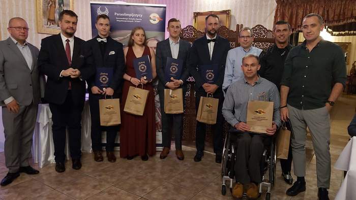 Pokaż zdjęcie: Uczetnicy spotkania stoją obok siebie w miejscu przeznaczonym dla fotoreporterów, jest zawodnik na wózku inwalidzkim, wielu trzyma okolicznościwowe torby prezentowe, są uśmiechnięci, jest pośród nich Dyrektor Oddziału Lubuskiego PFRON