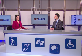 Pokaż zdjęcie: Szerokie ujęcie studia telewizyjnego programu "BEZ BARIER". Przy długim białym biurku siedzą dwie osoby: prowadząca w amarantowej marynarce oraz zaproszony gość w kraciastej marynarce. Przód biurka zdobią duże niebieskie ikony dostępności (osoba z laską, aparat słuchowy, język migowy, niepełnosprawność intelektualna, wózek inwalidzki). W tle widać ekrany z nazwą programu.