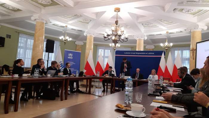 Pokaż zdjęcie: W sali konferencyjnej Urzędu Wojewódzkiego przy okrągłym stole siedzą kobiety i mężczyźni. Część z nich ma słuchawki na uszach. Za stołem prezydialnym stoją banery Lubelskiego Urzędu Wojewódzkiego w kolorze granatowym oraz roll-up UNHCR.
