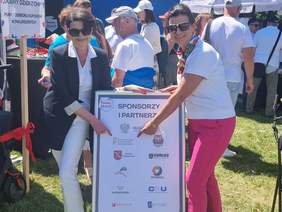 Pokaż zdjęcie: Dwie uśmiechnięte kobiety stoją na świeżym powietrzu podczas wydarzenia plenerowego. Obie są elegancko ubrane – kobieta po lewej ma ciemną marynarkę i białe spodnie, a kobieta po prawej białą koszulkę i różowe spodnie. Obie trzymają dużą tablicę z logiem OHP i napisem „Sponsorzy i partnerzy” oraz logotypami różnych instytucji, w tym PFRON. W tle widać tłum ludzi i białe namioty targowe. Po lewej stronie znajduje się plansza z napisem „Dobry Dzień z OHP – Punkt odbioru kuponów konkursowych”.