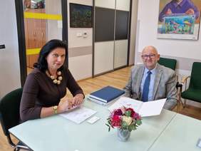 Pokaż zdjęcie: Kobieta i mężczyzna elegancko ubrani siedzą przy stole. Rozmawiają pochyleni nad dokumentami.