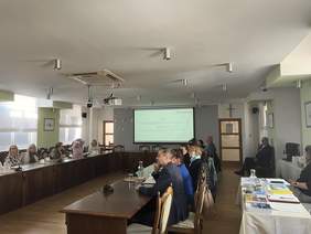 Pokaż zdjęcie: Grupa osób przy stole w kształcie litery U podczas prezentacji w jasnej sali konferencyjnej. Uczestnicy patrzą na ekran projekcyjny; na stołach widoczne mikrofony, woda i materiały drukowane.